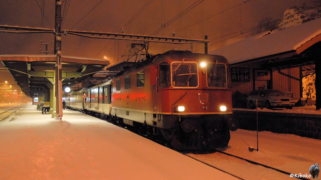 Das Bild zeigt eine rote Elektrolokomotive bei einen Halt am beleuchteten Bahnsteig im Schneetreiben.