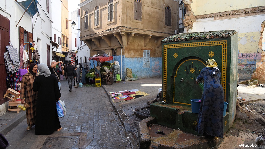Das Bild zeigt einen kleinen Platz. Rechts sind an einem grünen Steinblock zwei Wasserhähne. Eine Frau im Dunkelblauen Mantel füllt zwei blaue Wassereimer. Rechts unterhalten sich zwei Frauen in der Gasse. Im Hintergrund sind in der Gasse geöffnete Läden und waren vor den Eingängen zu sehen.