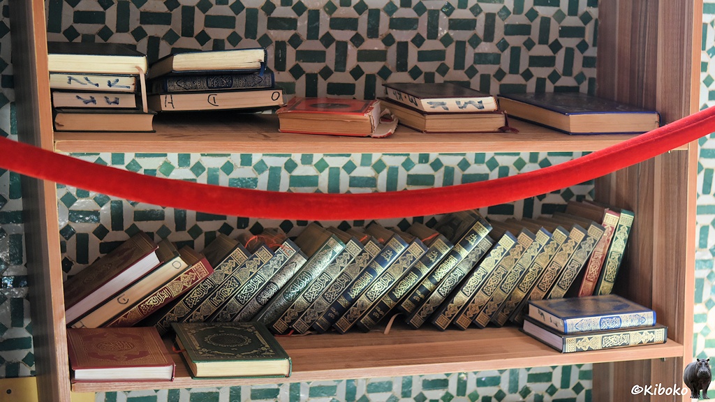 Das Bild zeigt ein Holzregal mit 36 Büchern auf zwei Regalböden. Die Bücher sind grün, blau oder rot eingefasst und sind mit goldenen Mustern geprägt.
