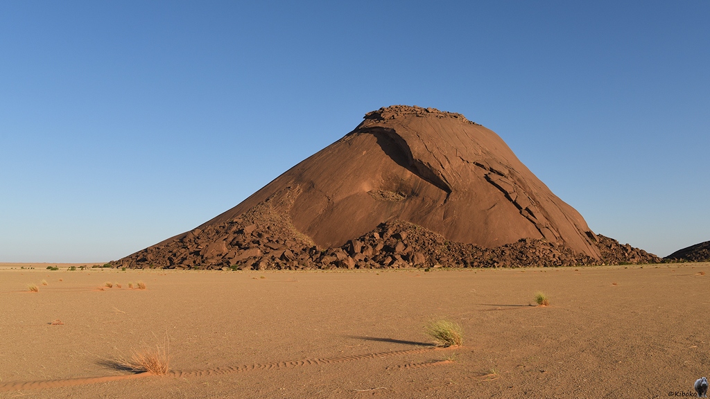 Das Bild zeigt einen Berg aus rotbraunem Fels im Morgenlicht. Über den Berg zieht sich eine Stufe im sonst glatten Fels. Um den Gipfel und rund um den Berg liegen Felsbrocken.