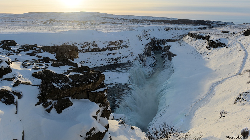 Das Bild zeigt die untere Stufe des Wasserfalls von der Seite. Der Wasserfall stürzt in eine tiefe Schlucht. Auf der gegenüberliegenden Seite bedeckt ein Eispanzer die Felsen. Im Vordergrund ist am linken Rand eine Felsformation, die den Blick auf die obere Stufe versperrt. Im Hintergrund ist eine flache Hügelkette. Darüber ist die tiefstehende Sonne im Bild.