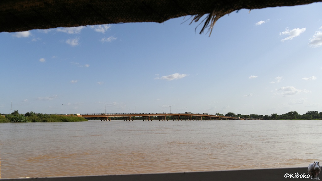 Das Bild zeigt eine braune Betonbrücke mit 11 Pfeilern die in einen Bogen knapp über den Hochwasser führenden Fluss geht. Oben und Unten ragen Bordwand und Dach ins Bild.