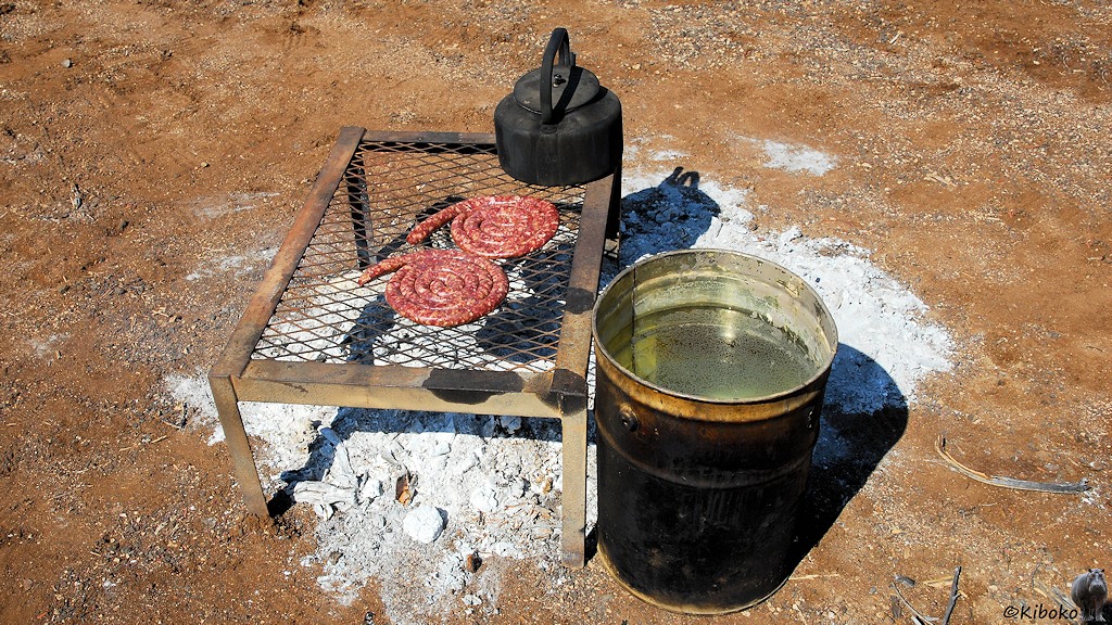Ein Gestell mit einem Grillrost steht über dem Lagerfeuer. Auf dem Grill sind zwei Wurstschnecken und ein schwarzer Teekessel. Neben dem Grill steht eine Wassertonne am Feuer.