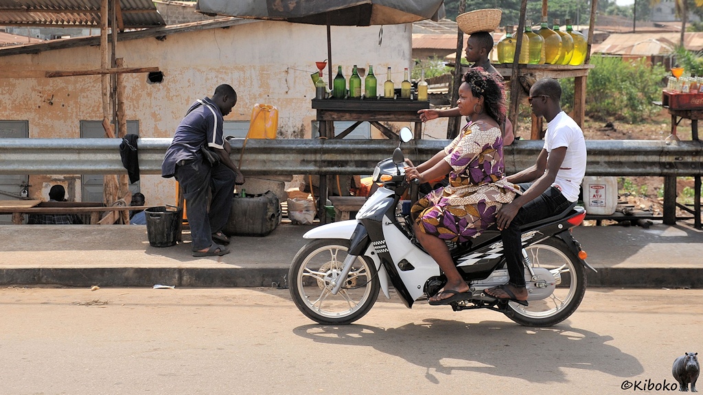 Eine Dame mit dicken Hinern fährt Motorrad. Ich Beifahrer hält sich an ihrem Hintern fest. Im Hintergrund wird Benzin umgefüllt.