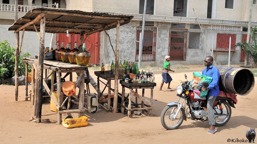 Ein Motorradfahrer hält an einer Tankstelle mit Flaschen auf einem Holzgerüst. Auf dem Gepäckträger hat er ein offenes 200-liter Fass.