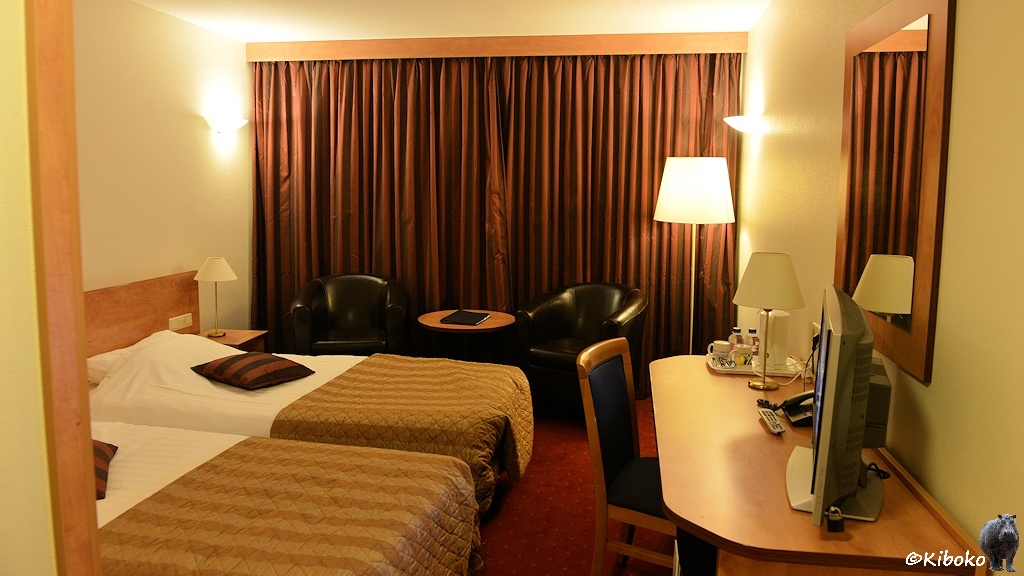 Das Bild zeigt ein Hotelzimmer mit zwei Einzelbetten, zwei schwarzen Schalensesseln und einem Sideboard mit Fernseher. Die braunen Gardinen sind zugezogen.