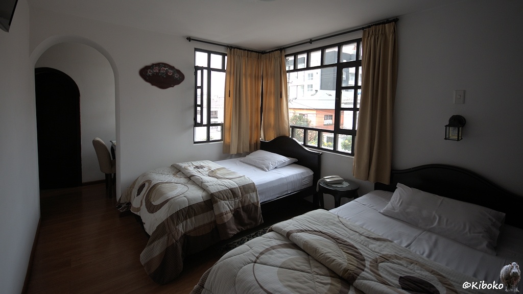 Das Bild zeigt ein Hotelzimmer mit einem Einzel- und einem Doppelbett. Es hat ein Eckfenster. Hinter dem Bogen am rechten Bildrand ist ein Schreibtisch und der Zugang zum Badezimmer.