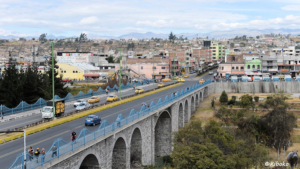 Das Bild zeigt eine Bogenbrücke mit 10 Bögen aus hellgrauen Steinen über ein Tal. Auf der Brücke ist eine vierspurige Straße mit vielen gelben Taxis. Die Geländer sind blau gestrichen. Im Hintegrund ist eine Stadt.