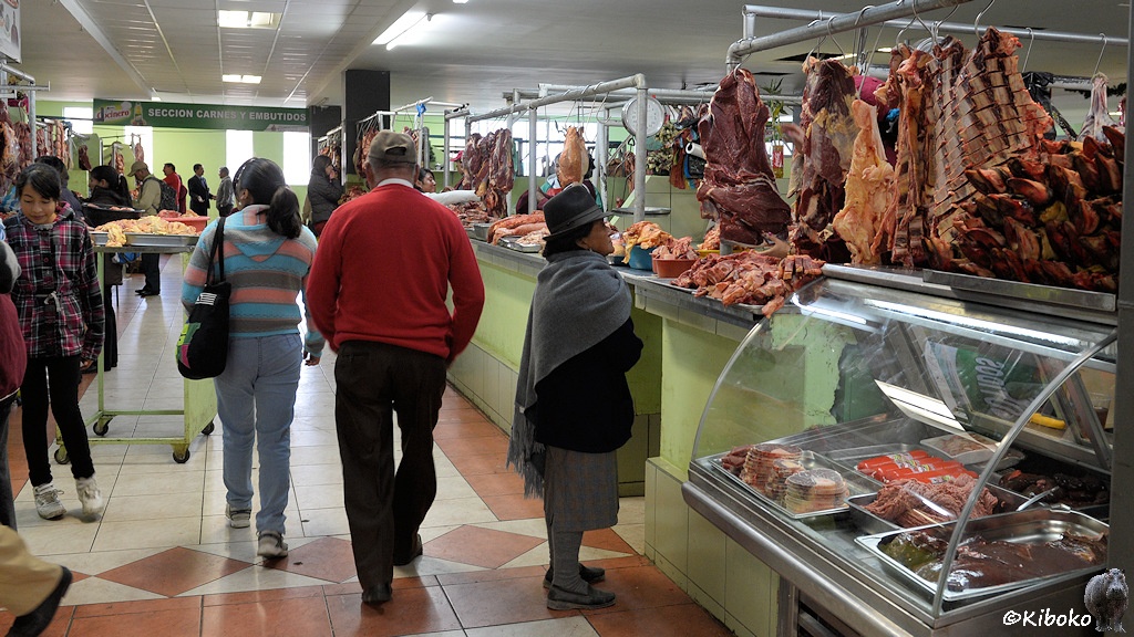 Das Bild zeigt hellgrüne Verkaufsstände mit Fleischwaren in einem geschlossenen Markt. Mehrere Leute schlendern auf beigen Fliesen durch die Gänge. Ein Frau mit grauem Poncho und grauem Hut schaut sich den rechten Verkaufsstand an.