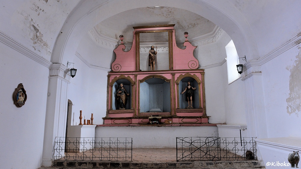 Das Bild zeigt die Innenansicht mit dem ehemaligen Altarraum. Der Putz bröselt von den weißen Wänden. Hinten sind Reste des Hochaltars als rosa lackierte Holzkonstruktion. Unten ist Platz für drei Figuren. Die mittlere Figur in einer hellblauen Nische fehlt. Oben ist in der Mitte eine weitere Figur mit einem Hirtenstab.