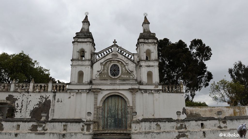 Das Bild zeigt das Portal einer weißen Kirche mit abblätternder Farbe. Die hellblaue Holztür ist verwittert. Über der Tür ist ein Innschrift um ein ovales Fenster: Santuario Miro Flores - La Maria. Daneben sind zwei kleine Türmchen mit jeweils einer Glocke.