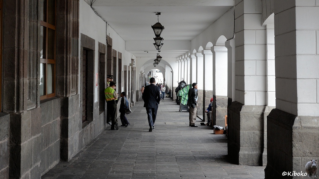 Das Bild zeigt einen Arkardengang. Decke, Wände und Säulen sind oben weiß. Dre Boden ist aus grauem Naturstein. Eine Polizistin steht am rechten Bildrand, während zwischen den Säulen die Schuhputzer arbeiten.