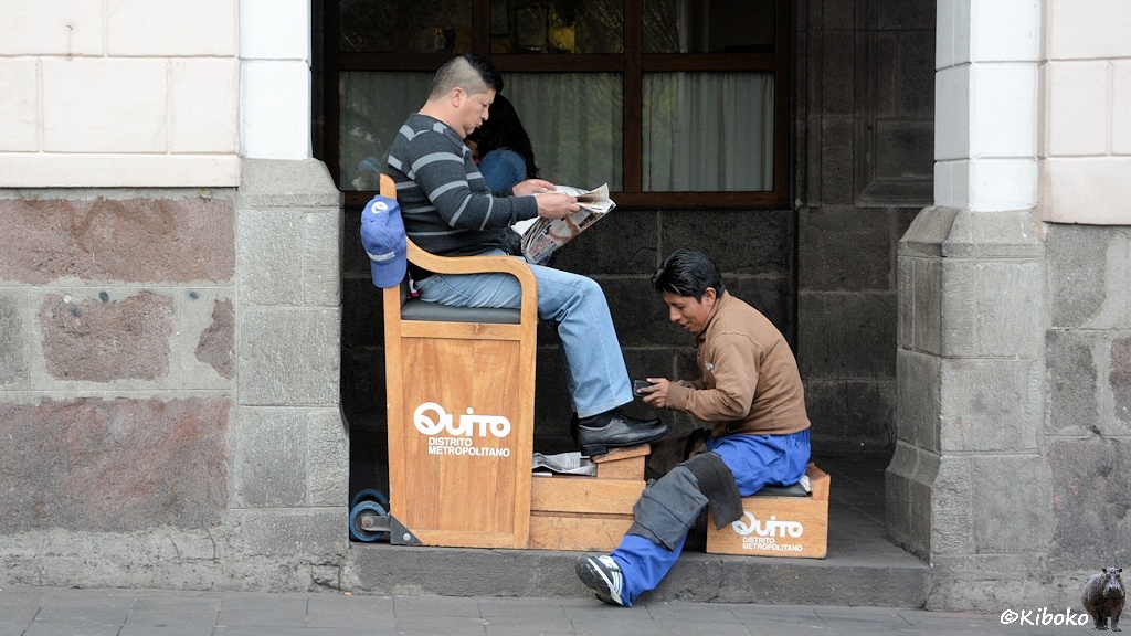 Das Bild zeigt einen Schuhputher bei der Arbeit. Er sitzt auf einer Kiste während er einen zeitungslesenden Mann auf einem erhöhten mobilen Sitz die Schuhe putzt. Sitz und Kiste tragen die Aufschrift: Quito Disterito Metropolitano.