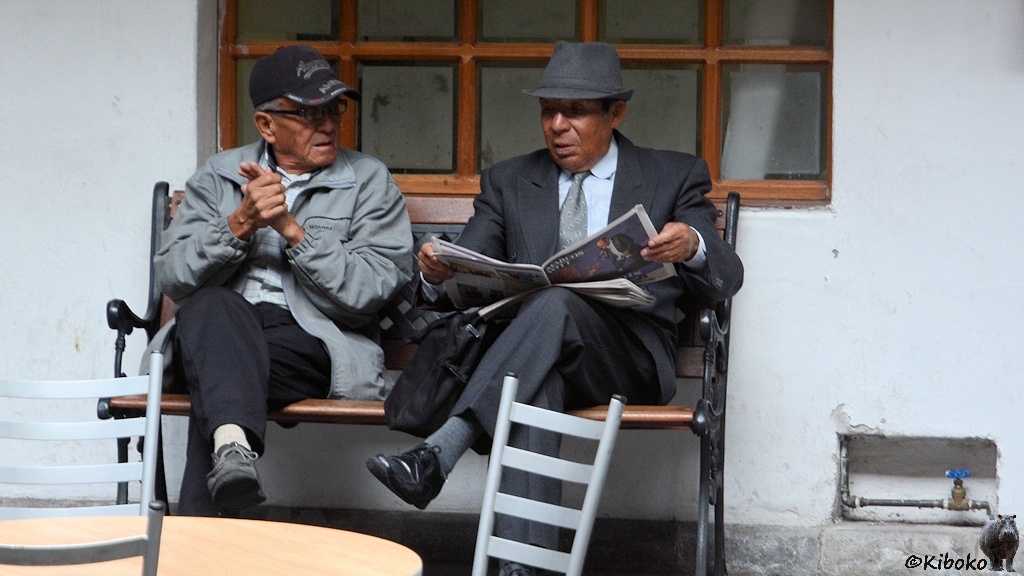 Das Bild zeigt zwei ältere Männer auf einer Bank. Der rechte Mann trägt einen dunkelgrauen Anzug mit Hemd, Krawatte und Hut. Er liest Zeitung. Während der linke Mann leger gekleidet ist und eine dunkle Baseballkappe trägt.