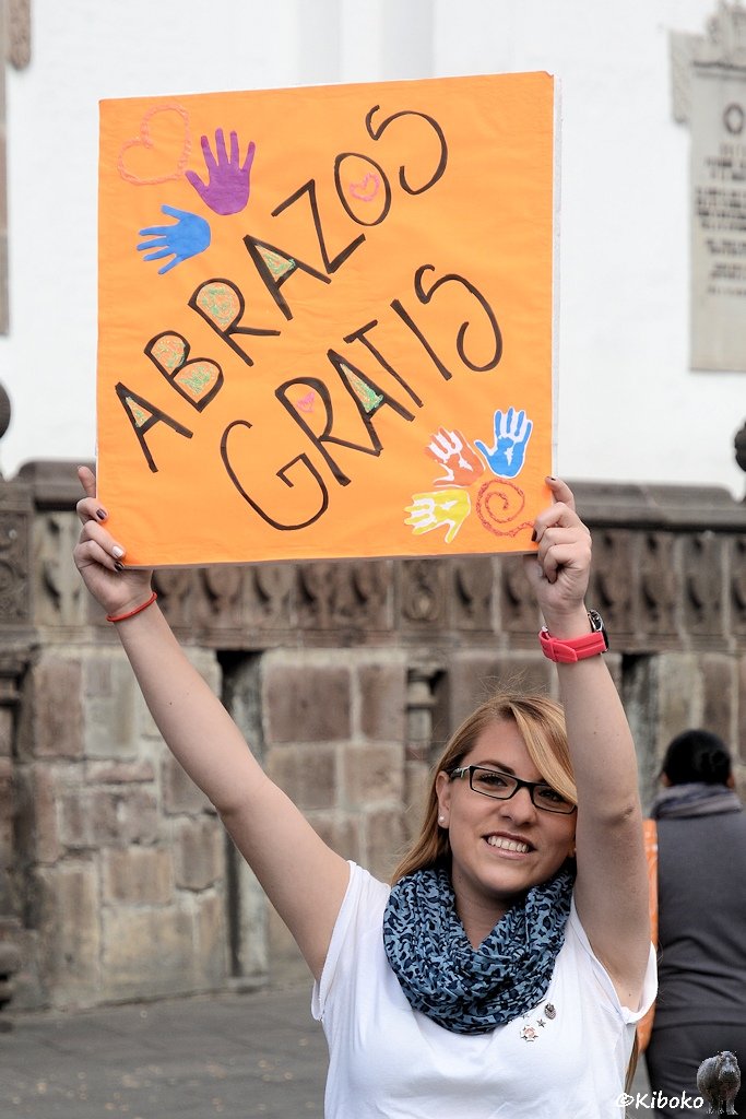 Das Bild zeigt eine Hochformataufnahme einer jungen blonden Frau im weißen T-Shirt die ein oranges Schild hochhält. Darauf stet in schwarzer Schrift: Abrazos Gratis.