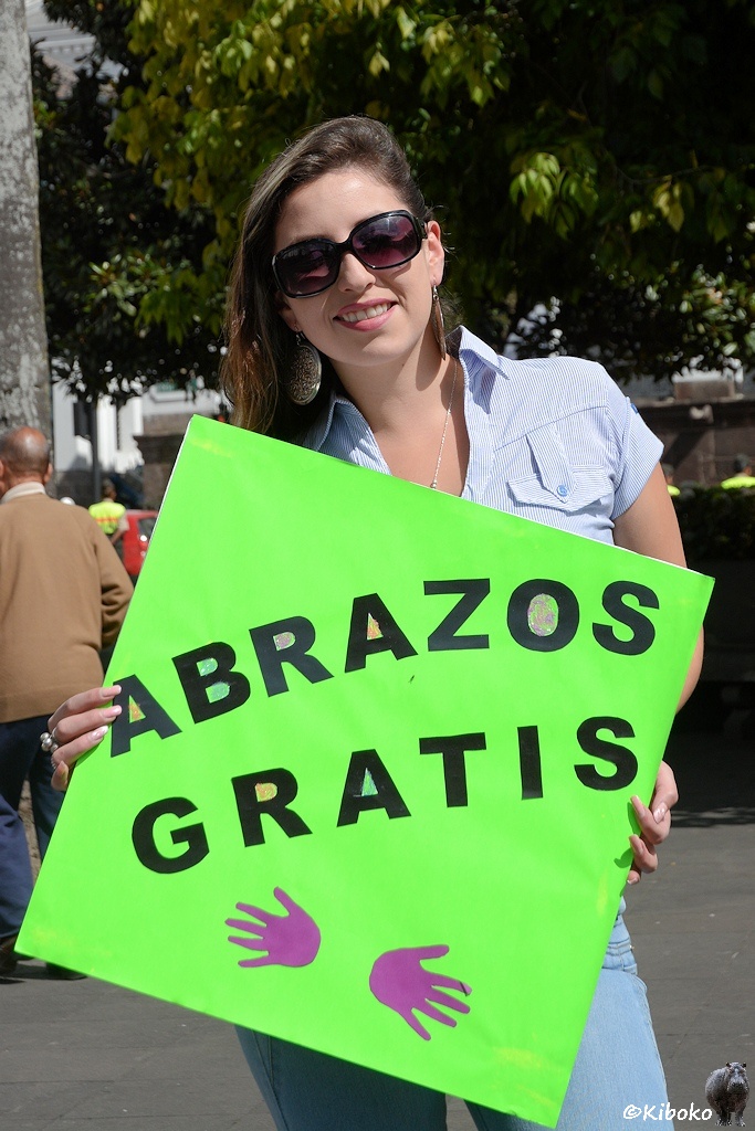 Das Bild zeigt eine junge lächelnde Frau mit dunklen Haaren und hellblauer Bluse. Sie trägt ein großes grünes Schild mit schwarzer Schrift vor der Brust. Auch auf dem Schild seht Abrazos Gratis.