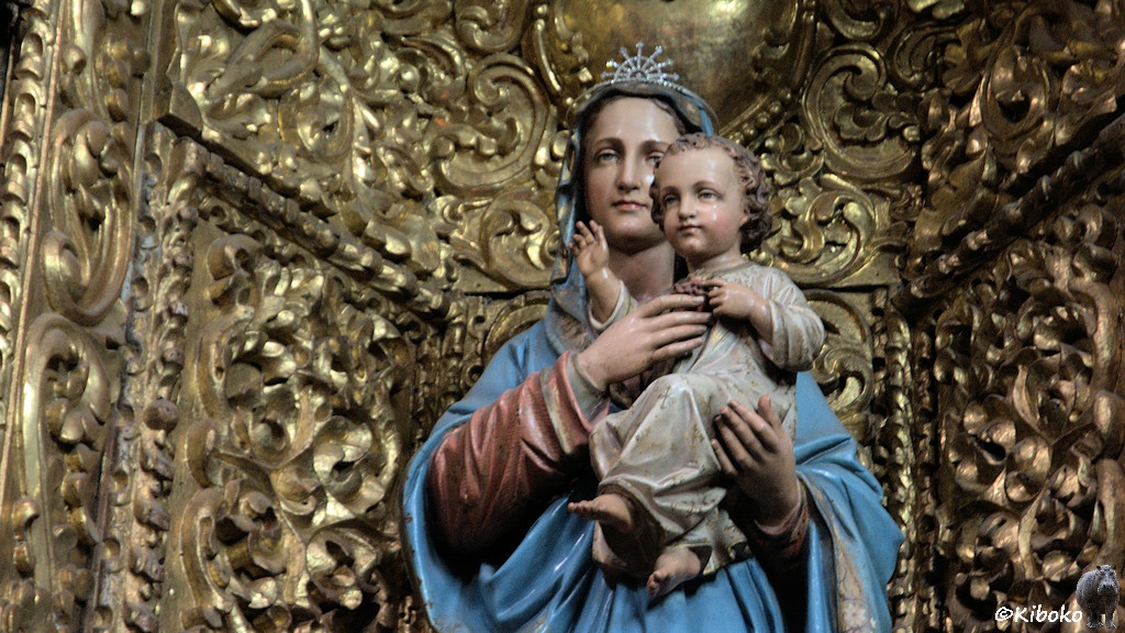 Das Bild zeigt das Porträt einer Marienstatur in einer hellblauen Robe mit einem kleinen Kind auf dem Arm. Der Hintergrund sind vergoldete Schnitzereien.