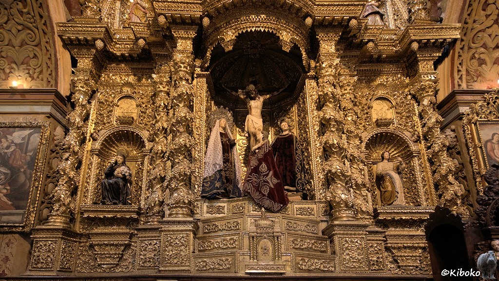 Das Bild zeigt einen Altar mit drei Nischen. In der Mitttleren ist ein Kruzefix mit drei weiteren Figuren. Bis auf die Figuren ist alles mit Gold belegt.