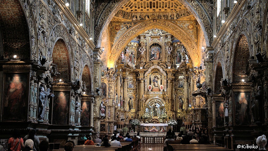 Das Bild zeigt das innere einer Kirche. Die Seiten, Decken und insbesondere der Altarraum sind mit Figuren, Reliefs verziert und mit Gold belegt. Am unteren Bildrand sitzen Kirchenbesucher, die gerade die Messe besucht haben.