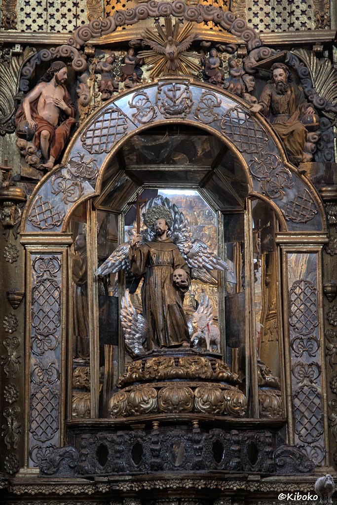Das Bild zeigt einen Mann mit Mönchskutte, der in der rechten Hand ein Kreuz hochhält und in der linken Hand einen Schädel hält. Die Nische ist mit Figuren und gold belegten Reliefs verziert.