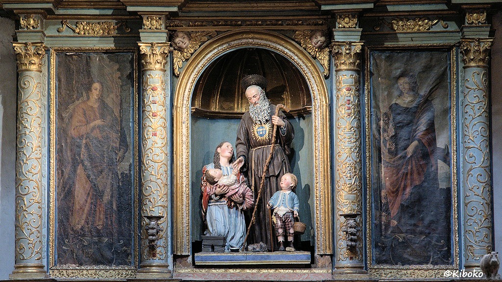 Das Bild zeigt eine sitzende Frau mit Kind neben einem Hirten der nach der Hand des Kindes greift. Ein weiteres kleines Kind steht vor dem Hirten. Auf beiden Seiten sind Gemälde.
