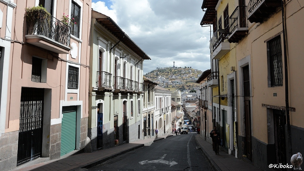 Das Bild zeigt eine schmale talwärts führende Straße. Auf beiden Seiten ist ein schmaler Gehweg. Zweistöckige Häuser mit Balkonen im Obergeschoss säumen die Straße. Im Hintergrund ist ein Berg mit einer Statue an der Spitze.