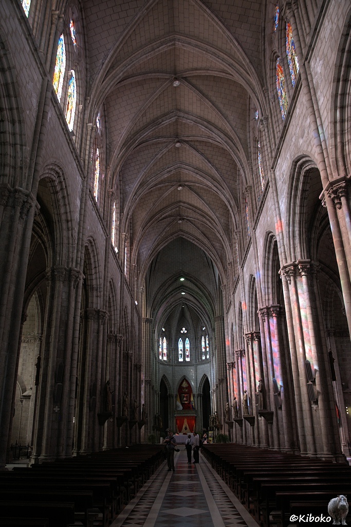 Das Bild zeigt eine Hochformataufnahme vom Mittelschiff in Richtung Altar. Der hohe Innenraum ist sehr schlicht gestaltet aus graubraunen Steinen. Auf der rechten Seite sind die Säulen rot blau und gelb gemustert, da die Sonne durch bunte Fenster schein.