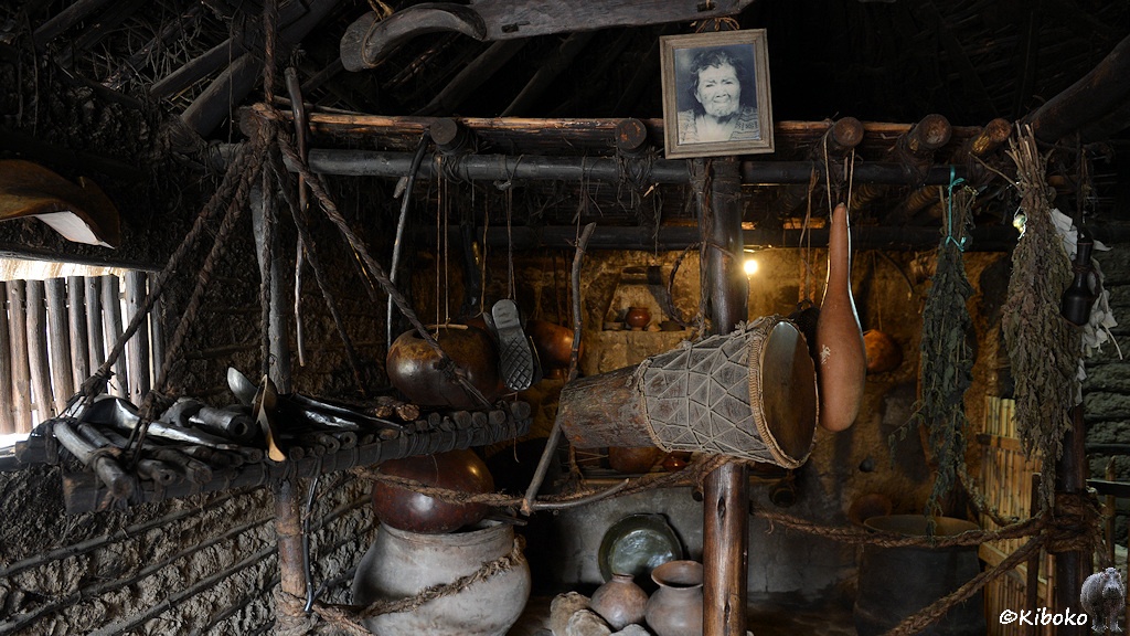 Das Bild zeigt das innere eines Steinhauses mit vielen Werkzeugen und Haushaltsgeräten, die an einer Stange aufgehängt ist. Oben hängt gibt es ein gerahmtes Bild der Bewohnerin.