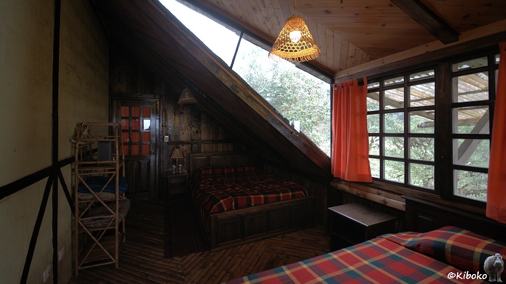 Das Bild zeigt die Innenansicht einer Dachkammr. Links ist eine Fachwerkwand. Rechts ist die Dachschräge. Ein Bett steht unter der Dachschräge. Am rechten Bildrand ist ein Sprossenfenster. Ein weiteres Bett mit rot-gelb-blau karrierter Decke steht vor dem Fenster.