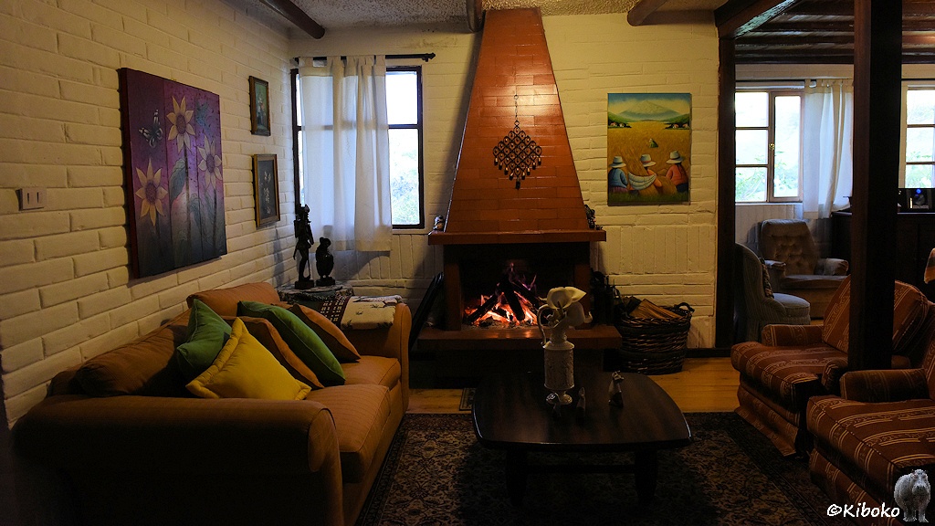 Das Bild zeigt die Innenansicht eines Raumes. Hinten ist ein Kamin, indem ein Feuer brennt. Links steht ein Sofa. Rechts stehen wei Sessel.