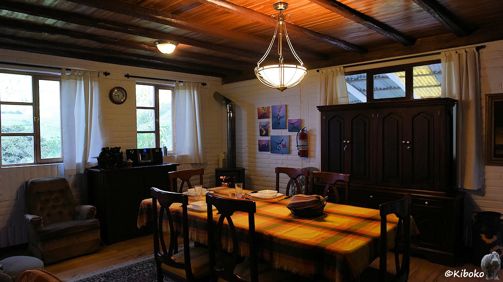 Das Bild zeigt ein Esszimmer. In der Mitte steht ein Tisch mit gelber Tischdecke und 6 Stühlen. Im Hintergrund sind zwei Fenster, ein Schrank und ein Sessel. Eine Hängeleuchte beleuchtet den Tisch.