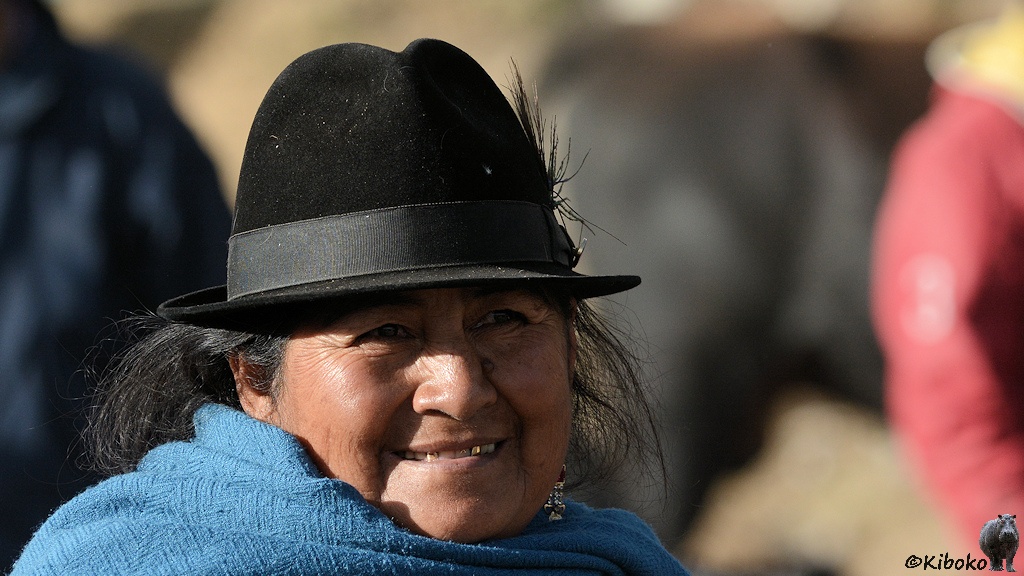 Das Bild zeigt ein Porträt einer Frau mit schwarzem Hut und blauem Umhang. Sie lächelt und die Goldzähne blitzen hervor.