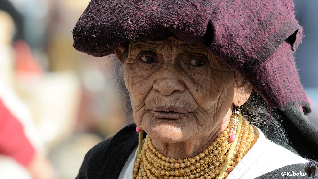 Das Bild zeigt ältere Frau mit sehr vielen Falten im Gesicht. Sie trägt ein gefaltetes violettschwarzes Tuch auf dem Kopf und eine Kette mit goldenen Kugeln.