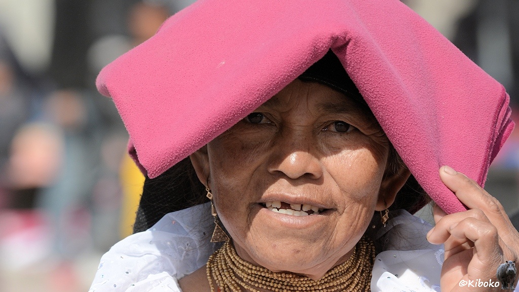 Das Bild zeigt ein Porträt einer lächelnden Frau. Sie trägt ein dunkelrosa Kopftuch. Das Kopftuch ist wie eine Decke. Sie ist mehrfach gefaltet und wird wie ein Hausdach auf dem Kopf getragen.