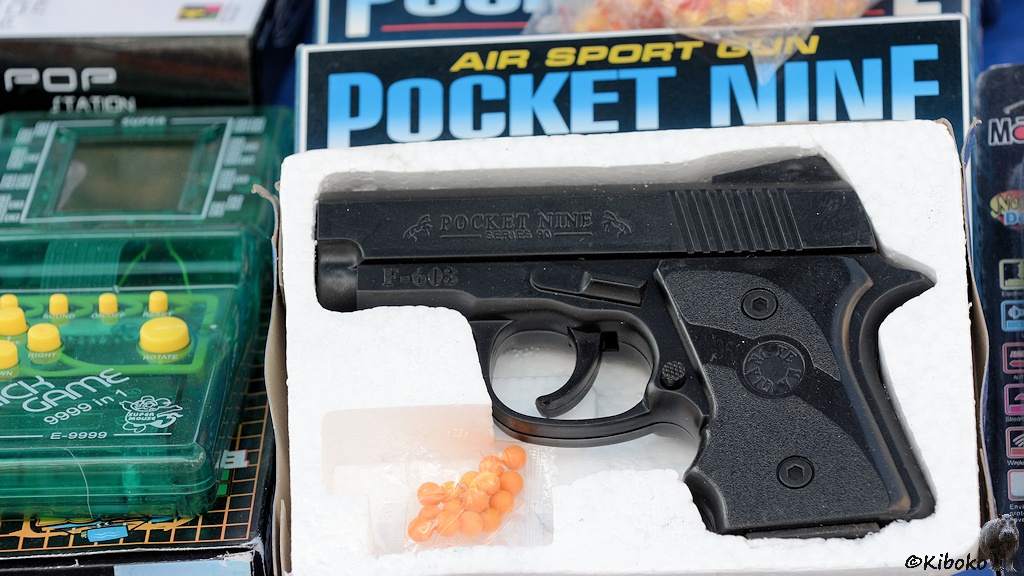 Das Bild zeigt eine schwarze Spielzeugplastikpistole in einer Styroporschachtel. Dabei liegt ein durchsichtiges Tütchen mit gelborangen Kugeln.