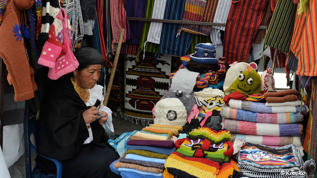 Das Bild zeigt eine alte Frau in weißer Bluse, schwarzen Rock und schwarzem Kopftuch auf einen Stand mit Stoffen. Sie häkelt gerade eine blaue Mütze. Zwischen den Stoffauslagen liegen fertige Gesichtermützen.