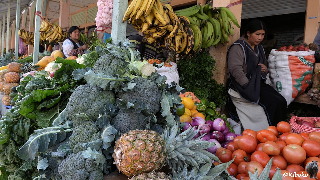 Das Bild zeigt einen Marktstand mit einen Berg Brokoli. Davor liegen Ananas. Dahinter liegen violette Zwiebeln und Tomaten. Darüber hängen grüne und gelbe Bananen. Eine Verkäuferin sitzt dezent im Hintergrund.