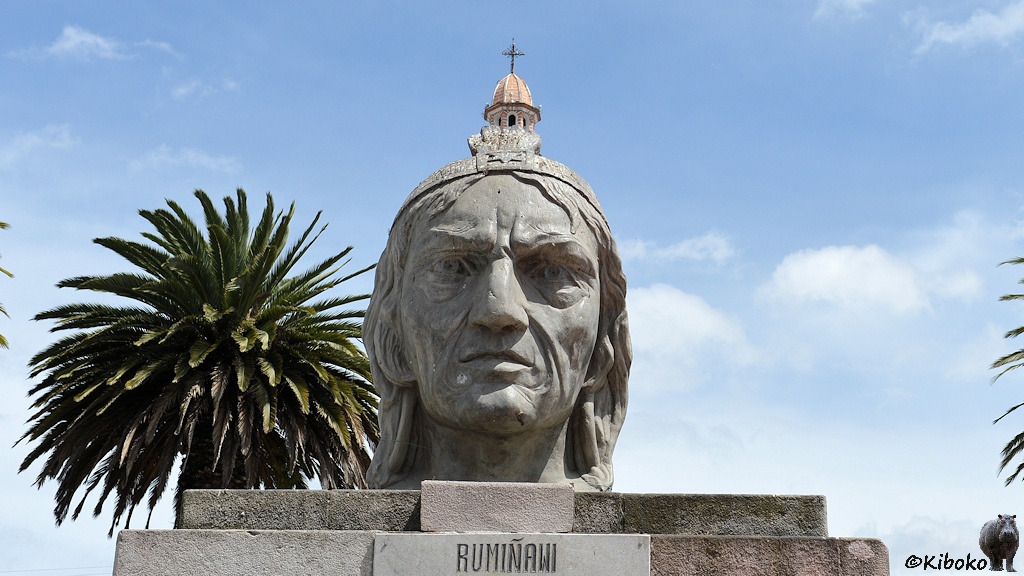 Das Bild zeigt einen großen Kopf aus grauem Stein eines Mannes mit langem Haar und Falten. Über dem Kopf ragt die Spitze eines Kirchturms mit einer runden Kuppel mit Kreus. Im Hintergrund ist links vom Kopf eine Palme.