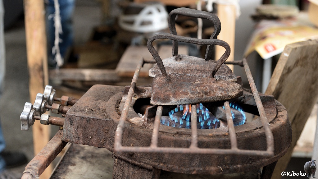 Das Bild zeigt einen stählernen Gasgrill mit mehreren Ringen mit blauen Flammen. Auf dem Rost stehen zwei rostige Bügeleisen mit großen Metallgriff.