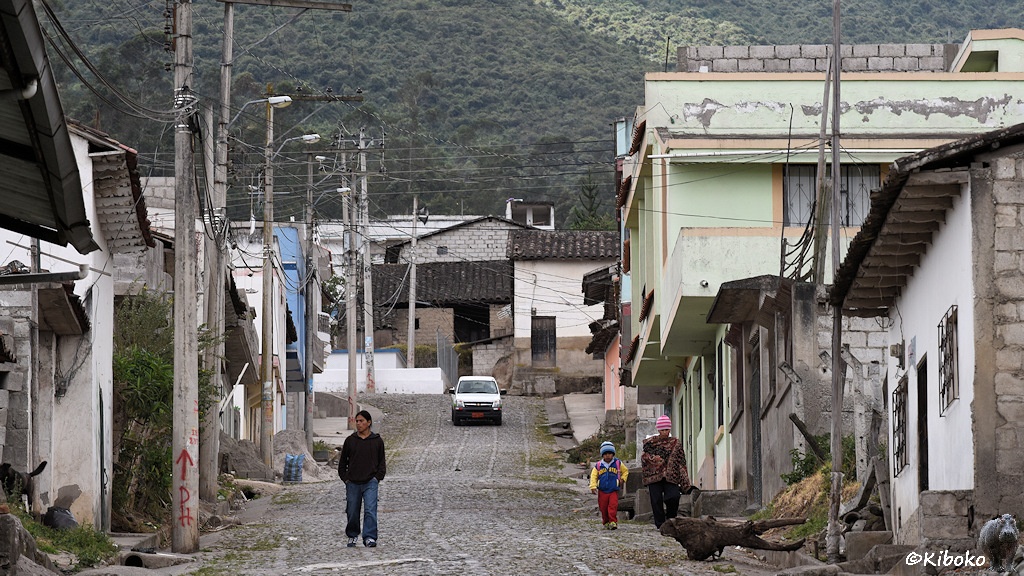 Das Bild zeigt eine bergauf führende Kopfsteinpflasterstraße mit einstöckigen Häusern. Betonmasten für Strom und Licht stehen am linken Straßenrand. Ein weißer Pickup und drei Personen kommen auf der Straße entgegen.