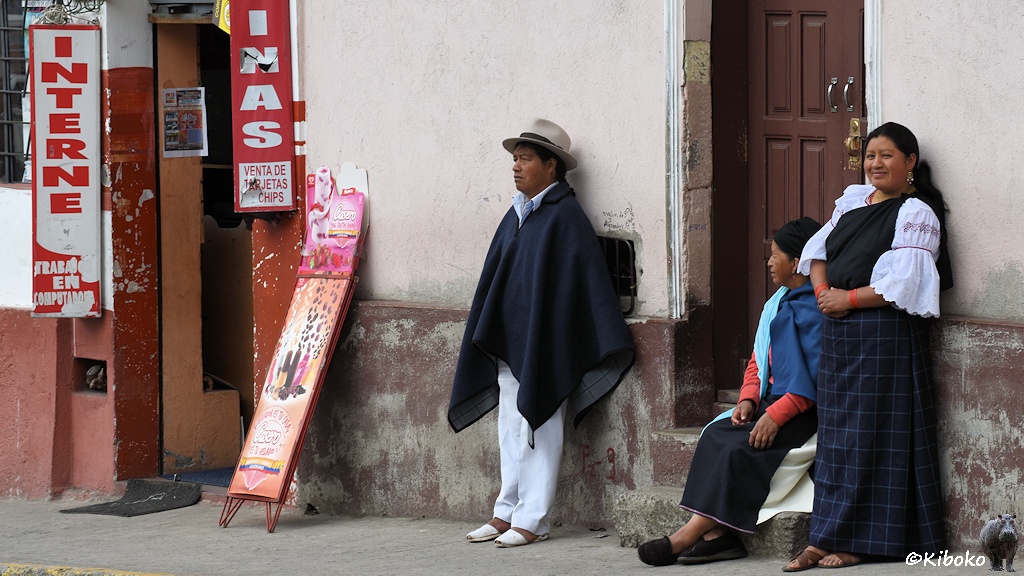 Das Bild zeigt zwei Frauen und einen Mann in traditioneller Kleidung an einer Hauswand. Eine Frau sitzt auf der Treppe einer Eingangstür. Links daneben ist ein Eingang in ein Geschäft mit einem Hinweisschild für Internet.