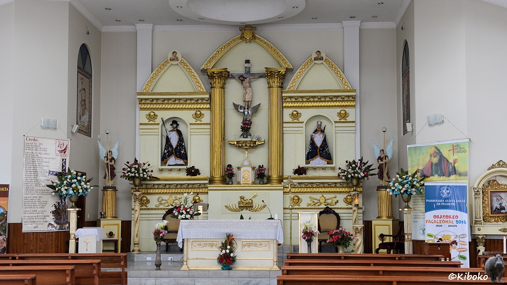 Das Bild zeigt den weißen Altar einer Kirche. Im Hintergrund wird der Jesus am Kreuz durch zwei goldene Säulen flankiert. Der Raum ist durch Blumen und kirchliche Werbeplakate geschmückt.
