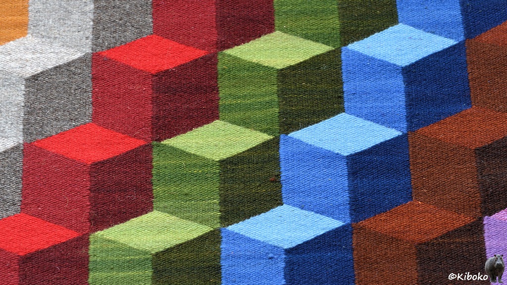 Das Bild zeigt einen Ausschnitt eines Wandteppichs. Hier sind diagonal aufsteigende Würfelmuster mit pseudo 3D-Effekt. Fünf Würfelreihen in grau,rot, grün, blau und braun laufen von links unten nach rechts oben durch das Bild.
