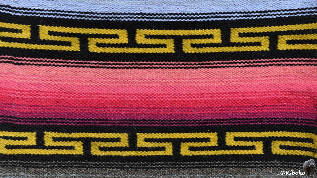 Das Bild zeigt einen Ausschnitt eines Wandteppichs. In der Mitte ist ein bewebter Streifen mit einem Übergang aus rosa bis nach violett.  Darüber und  darunter ist eine gelbe Linie auf schwarzem Grund. Die Linie ist eine Dreifachlinie mit schwarzen T-Einschnitten von oben und unten.