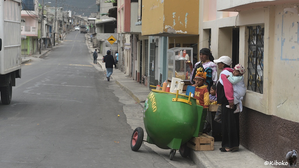 Das Bild zeigt eine gerade Teerstraße durch einen Ort. Am rechten Straßenrand steht eine grüne Bombenförmige Karre. Darauf stehen Plastikflaschen mit Soßen. Auf dem Gehweg stehen dahinter zwei Frauen mit drei Kindern.