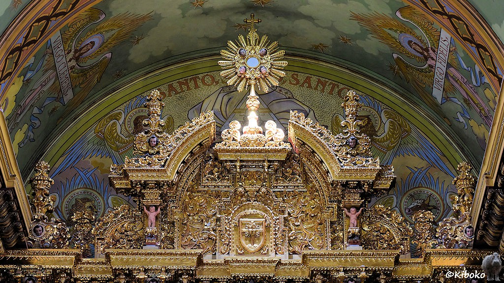 Das Bild zeigt die Spitze des goldenen Hochaltars. Die Spitze besteht aus einem Sonnenstern auf dem ein Kreuz steht. Darunter sind goldene Ornamente mit zwei Säulen. Die Säulen werden von zwei nackten Jungen getragen. Das Gewölbe darüber ist mit Gemälden verziert.