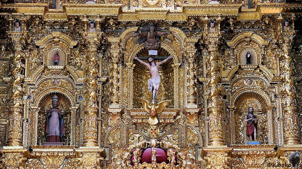 Das Bild zeigt einen Ausschnitt vom goldenen Hochaltar. In der Mitte ist Jesus am Kreuz in einer vergoldeten Niesche. Er schein auf einem goldenen Adler zu stehen. Auf beiden Seiten ist eine kleinere Figur in einer vergoldeten Niesche.