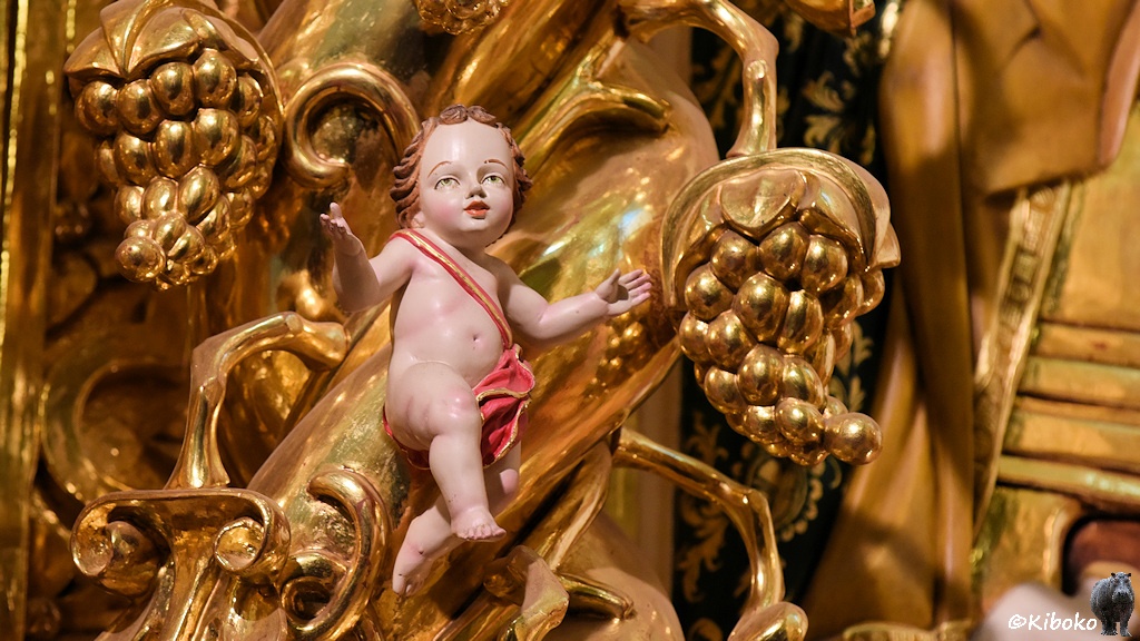 Das Bild zeigt zeigt einen nackten kleinen Jungen, der eine rote Windel mit Goldrand im Schritt hat, dass über ein rot-goldenes Band über die Schulter gehalten wird. Um die Figur sind goldene Ornamente. Zwei goldene Weinreben sind das Hauptmerkmal.