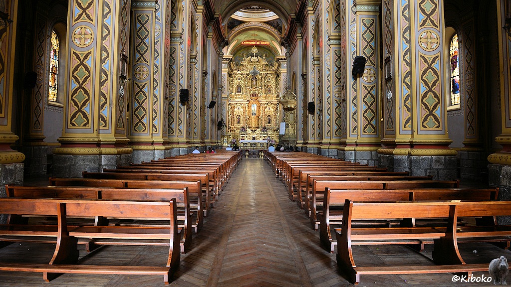 Das Bild zeigt das Mittelschiff einer Kirche vom Eingang her. Der Blick geht zwischen viereckigen, mit Farben dekorierten Säulen und Reihen mit Holzbänken auf einen gold glänzenden Hochaltar.