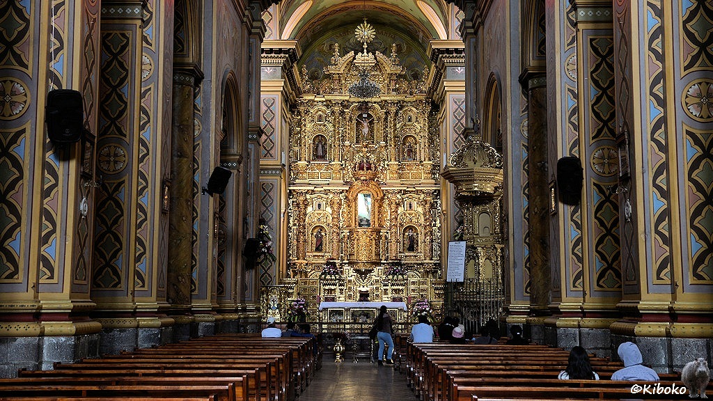 Das Bild zeigt das Innere einer Kirche mit dem vergoldeten Hochaltar in der Mitte. In den Holzbänken vor dem Altar sitzen ein paar Menschen. In der Mitte des Hochalars ist ein verspiegelter Glaskasten.